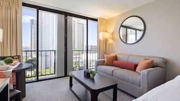 A modern hotel room with a balcony, a sofa, a coffee table, and a desk with a lamp.