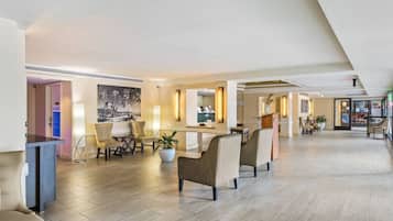 A spacious hotel lobby with a high ceiling, a reception desk, seating areas, and a large painting on the wall.