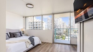 A modern bedroom with a large bed, a view of tall buildings, and a balcony with a railing. - Corner Bliss in the Heart of Waikiki 8F (Honolulu)