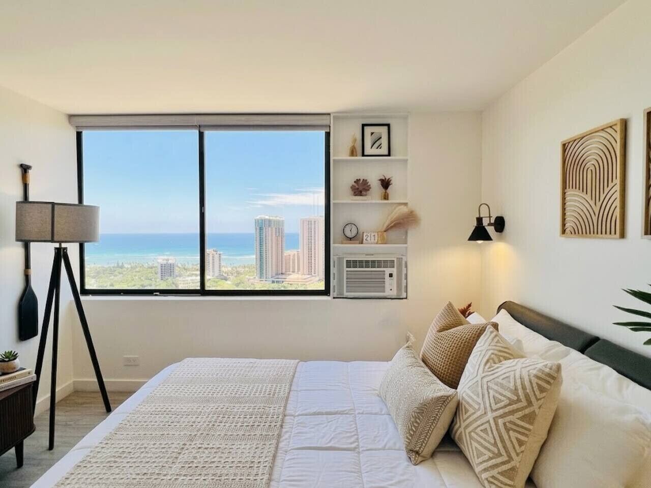 A bedroom with a large window, a bed, a bedside lamp, a wall clock, and a small shelf with decorative items.