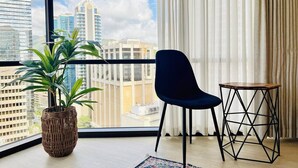 Une pièce avec une grande fenêtre, une plante en pot, un fauteuil sombre et une petite table en bois.