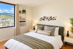 A bedroom with a bed, two bedside tables, a wall-mounted lamp, a decorative leaf wall art, a window with a view of buildings and mountains, and a shelf with a plant and a framed picture. - Stunning Mountain View Studio – High Floor Waikiki 3107B (Honolulu)