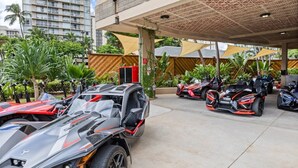 Een parkeerplaats met meerdere luxe voertuigen, waaronder een rode sportauto en een zilveren SUV, onder een overdekte constructie met een versierd plafond.