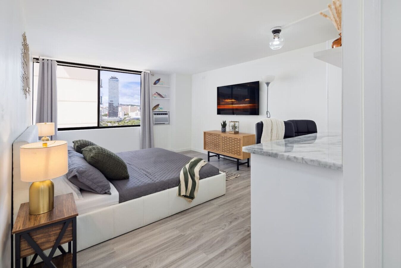 A modern bedroom with a bed, bedside table, lamp, TV, and a view of a cityscape through the window.