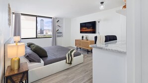 A modern bedroom with a bed, bedside table, lamp, TV, and a view of a cityscape through the window. - Waikiki Tranquil Retreat (Honolulu)