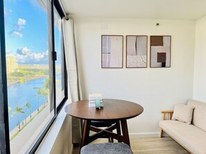 Una habitación con una mesa redonda de madera, un sofá y una ventana que da a una vista del río y edificios. Hay tres obras de arte enmarcadas en la pared.