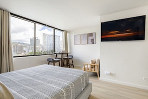 Habitación de hotel con una cama grande, un escritorio con dos sillas, un televisor en la pared y una ventana con vistas a la ciudad.