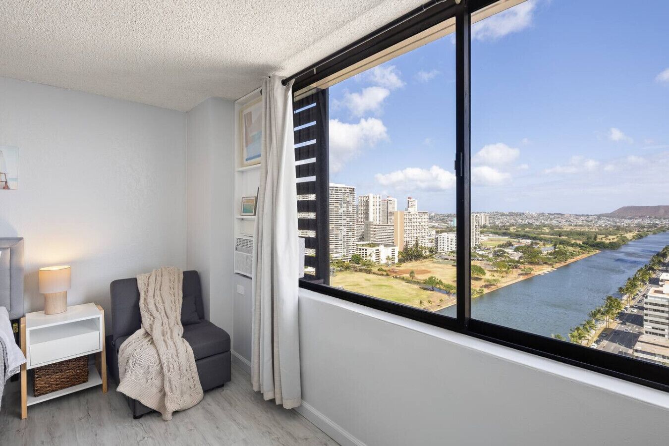 A modern room with a large window, a chair, a bedside table, and a lamp.