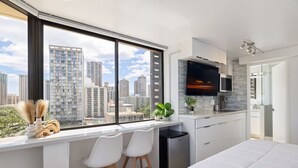 Un dormitorio moderno con un ventanal grande, una cama, un escritorio con sillas y una zona de cocina.