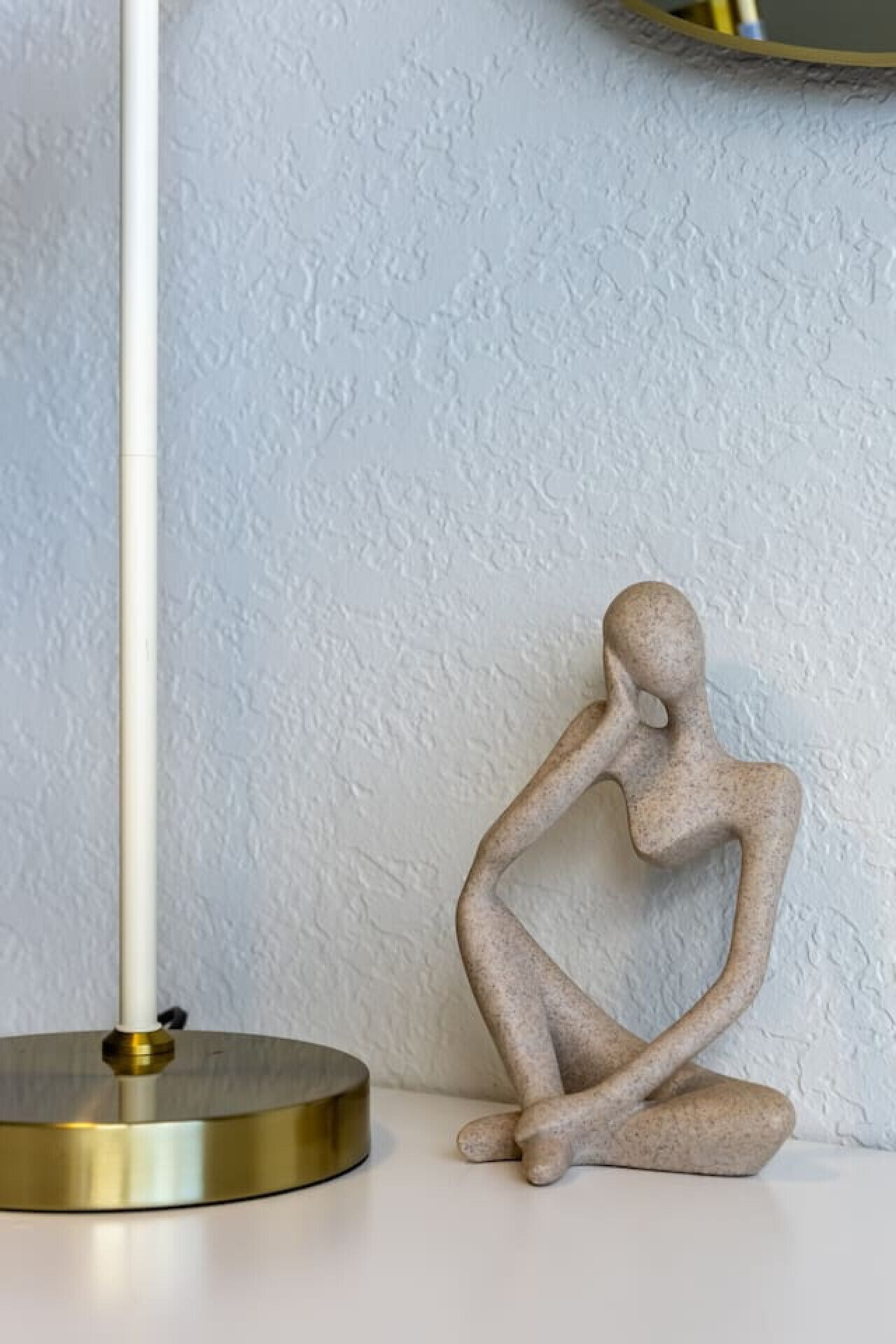 A minimalist sculpture of a seated figure next to a modern floor lamp on a white surface against a textured wall.