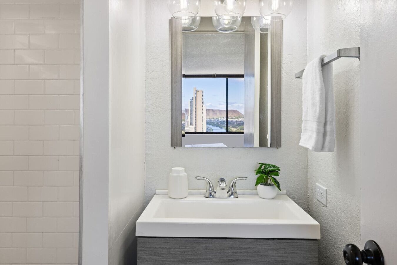 Un baño moderno con un lavamanos blanco, un espejo encima y vista a una ciudad a través de la ventana.