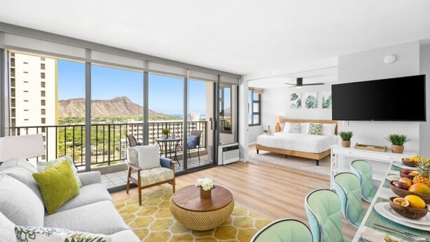 Una habitación de hotel moderna con balcón, televisor grande y zona de comedor con fruta.