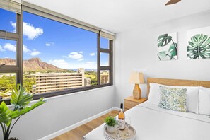 A hotel room with a large window, a bed, a bedside table with a lamp, and a tray with a bottle and glasses. - Waikiki Paradise w/ Ocean View & a Free Parking (Honolulu)