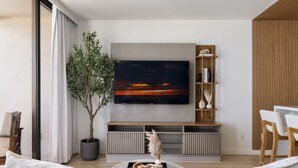 Ein modernes Wohnzimmer mit einem großen Flachbildfernseher an der Wand, einer Zimmerpflanze, einem Holzregal und einem runden Couchtisch mit Dekoration.