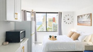 A modern hotel room with a bed, a desk with a microwave, a large window with a city view, a wall clock, and a framed picture. - Waikiki Skyline Corner Ocean View Studio (Honolulu)