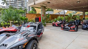 A parking area with several off-road vehicles, a covered structure with a patterned ceiling, and a backdrop of a tall building and lush greenery. - Waikiki Skyline Corner Ocean View Studio (Honolulu)