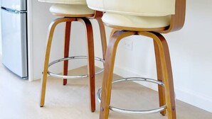 Ett modernt kök med två vita barstolar med träben, ett kylskåp i rostfritt stål och vita skåp.