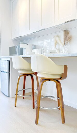Eine moderne Küche mit zwei weißen Barhocker mit Holzbeinen, einem Edelstahlkühlschrank und weißen Schränken.