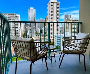 Ein Balkon mit zwei Korbstühlen und einem kleinen runden Tisch, mit Blick auf eine Stadtlandschaft mit hohen Gebäuden.
