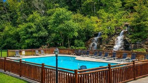 Une piscine rectangulaire entourée d’une clôture en bois, bordée d’arbres verdoyants et agrémentée d’une cascade.