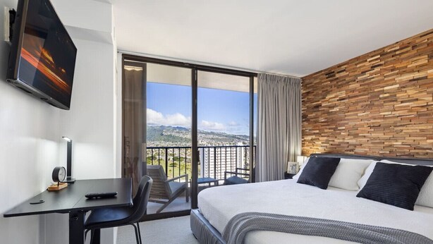 Une chambre d’hôtel avec un grand lit, un bureau avec une chaise, un téléviseur fixé au mur et un balcon offrant une vue sur la ville.