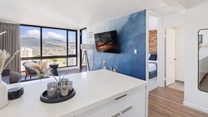 Une chambre d’hôtel moderne dotée d’une grande fenêtre, d’un téléviseur à écran plat fixé au mur, d’un bureau blanc sur lequel est posé un plateau de bougies, et offrant une vue sur le paysage urbain.