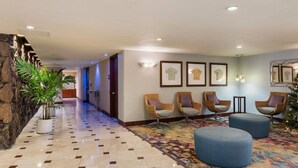 Un vestíbulo de hotel con pared de piedra, suelo de baldosas, sillas de mimbre, un otoman azul y obras de arte enmarcadas en las paredes.