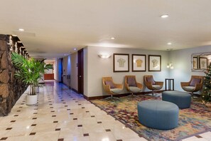 A hotel lobby with a stone wall, a tiled floor, wicker chairs, a blue ottoman, and framed artwork on the walls. - Waikiki Gem: Corner Unit with Ocean View 1 Parking (Honolulu)