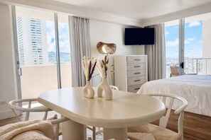 A hotel room with a balcony, a bed, a TV, a desk with chairs, and a dresser. - Waikiki Gem: Corner Unit with Ocean View 1 Parking (Honolulu)
