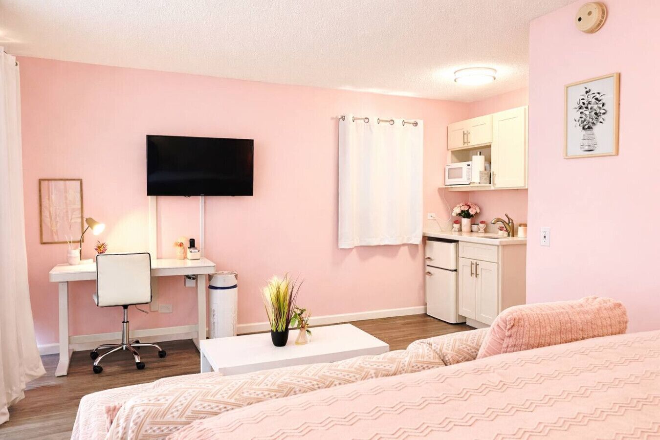 Een kamer in roze tinten met een bed, bureau, stoel, televisie en kitchenette.