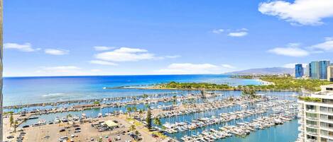 A marina with numerous boats docked, a parking area, and a view of the ocean and distant buildings.