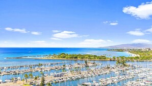 Una marina con numerosos barcos amarrados, un cielo azul despejado y una vista distante de la costa con vegetación y edificios.