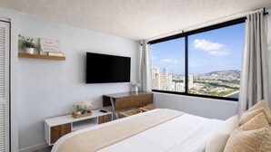 A modern bedroom with a bed, a TV mounted on the wall, a desk, and a view of a cityscape through a large window. - 27F Waikiki Cozy Studio w/ Great Ocean View B (Honolulu)