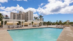 A swimming pool with lounge chairs and umbrellas, surrounded by tall buildings and palm trees. - 27F Waikiki Cozy Studio w/ Great Ocean View B (Honolulu)
