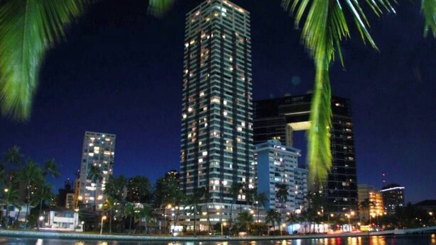 A nighttime cityscape with high-rise buildings, palm trees, and a body of water reflecting lights. - 27F Waikiki Cozy Studio w/ Great Ocean View B (Honolulu)