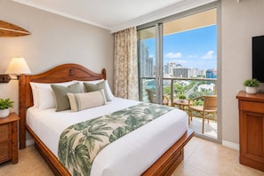A hotel room with a large bed, a balcony with a view of the city, a wooden headboard, and a small table with chairs. - Unobstructed Ocean View Spacious Studio (Honolulu)