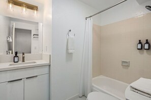 A bathroom with a white sink, mirror, shower curtain, and two bottles of shampoo on the wall. - Great Ocean Corner View 28F A (Honolulu)