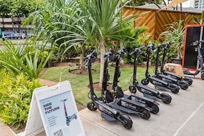 A row of electric scooters parked outdoors with a signboard nearby. - Waikiki Corner Panorama - 3107AB (Honolulu)