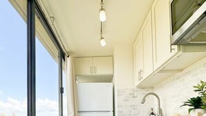 Une cuisine étroite avec des meubles blancs, un réfrigérateur en acier inoxydable, une fenêtre donnant sur des immeubles et un plancher en bois.
