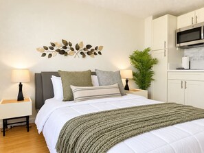 A bedroom with a bed, nightstands, a wall art piece, a potted plant, and a kitchenette area. - Waikiki Corner Panorama - 3107AB (Honolulu)