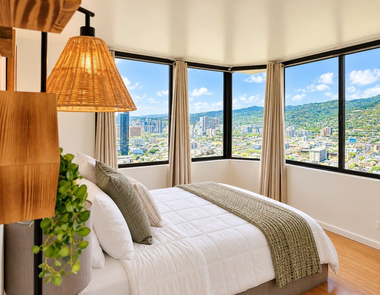 A bedroom with a large bed, a hanging lamp, and a view of a cityscape through large windows.