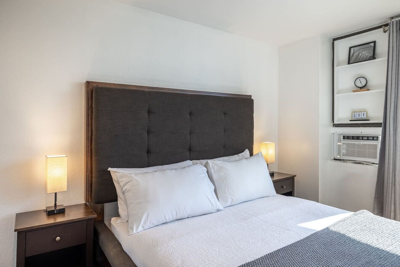 A hotel room with a large bed, two bedside tables with lamps, a wall-mounted shelf with a clock and a small photo frame, and a window with curtains.