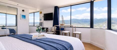 A modern hotel room with a large bed, a desk, a chair, a TV, and a view of the mountains.