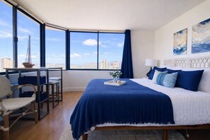 A hotel room with a large bed, a desk with a chair, and a view of the cityscape through large windows. - Rare Waikiki Corner 31F Unit with Stunning Views (Honolulu)