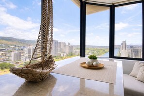 A modern room with a hanging chair, a round table with decorative items, and a large window offering a city view. - Rare Waikiki Corner 31F Unit with Stunning Views (Honolulu)