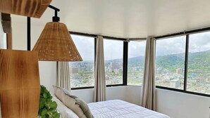 Een slaapkamer met een groot raam, een bed met wit beddengoed, een plant en een hanglamp.