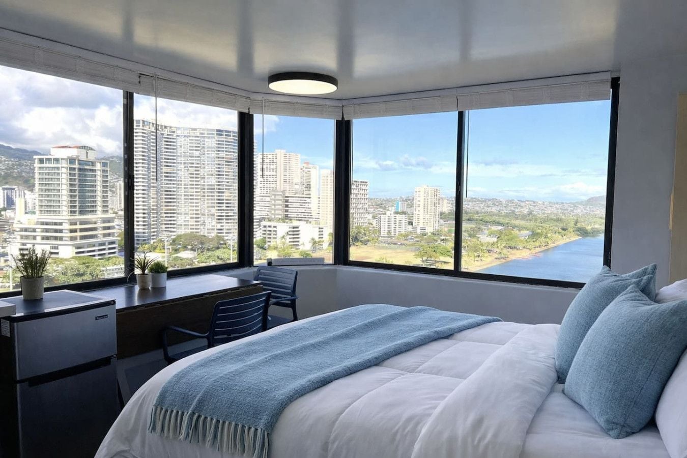 A bedroom with a large bed, a desk, and a chair, with a city view through large windows.