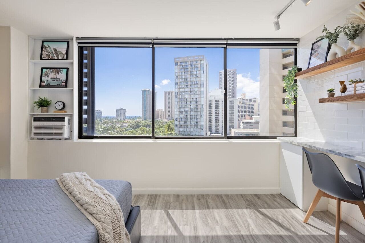 Un dormitorio moderno con una ventana grande, una cama, un escritorio y vistas a un paisaje urbano.