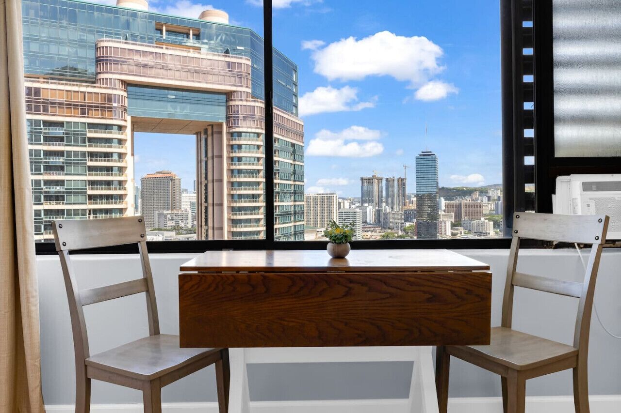 Una habitación moderna con un ventanal grande que da a una vista panorámica de la ciudad, con rascacielos y un cielo despejado.