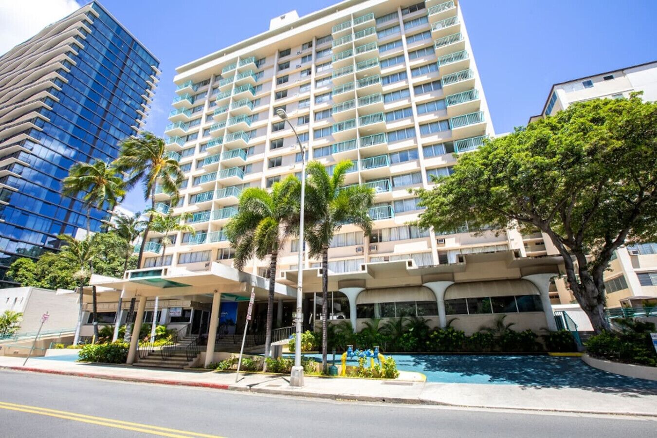 Un hotel de varios pisos con piscina, palmeras y un edificio moderno al fondo.